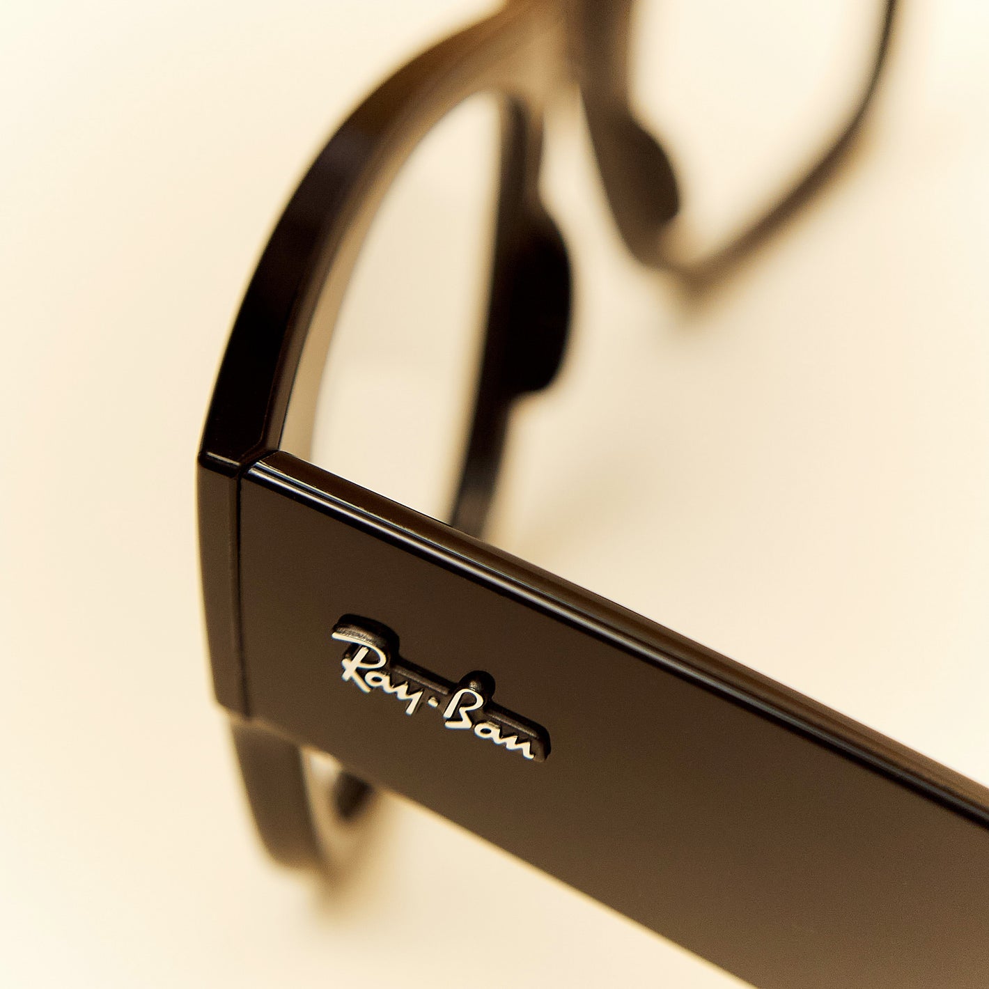 Close-up of a glossy black Ray-Ban eyeglass frame showing the silver Ray-Ban logo on the temple arm against a warm off-white background.