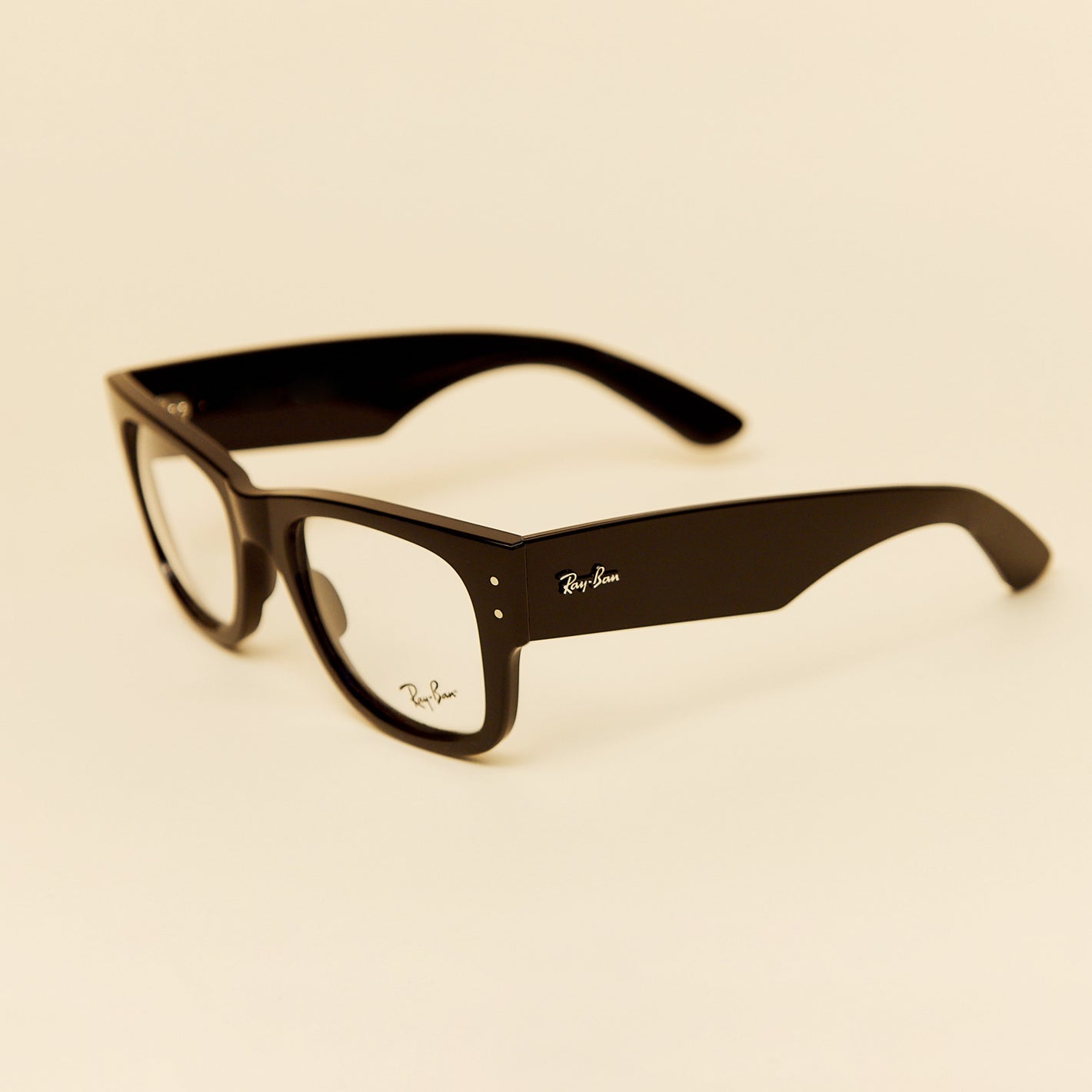 Ray-Ban Wayfarer-style eyeglasses with a glossy black frame and clear lenses, photographed on a warm cream background. The Ray-Ban logo is visible on the temple.