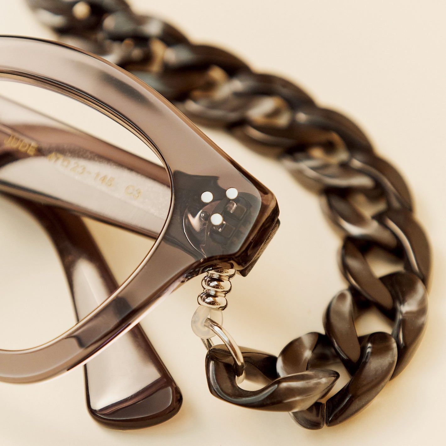 Close-up of a pair of translucent dark grey eyeglass frames with a matching chunky chain strap attached by a small metal ring, on a warm cream background.